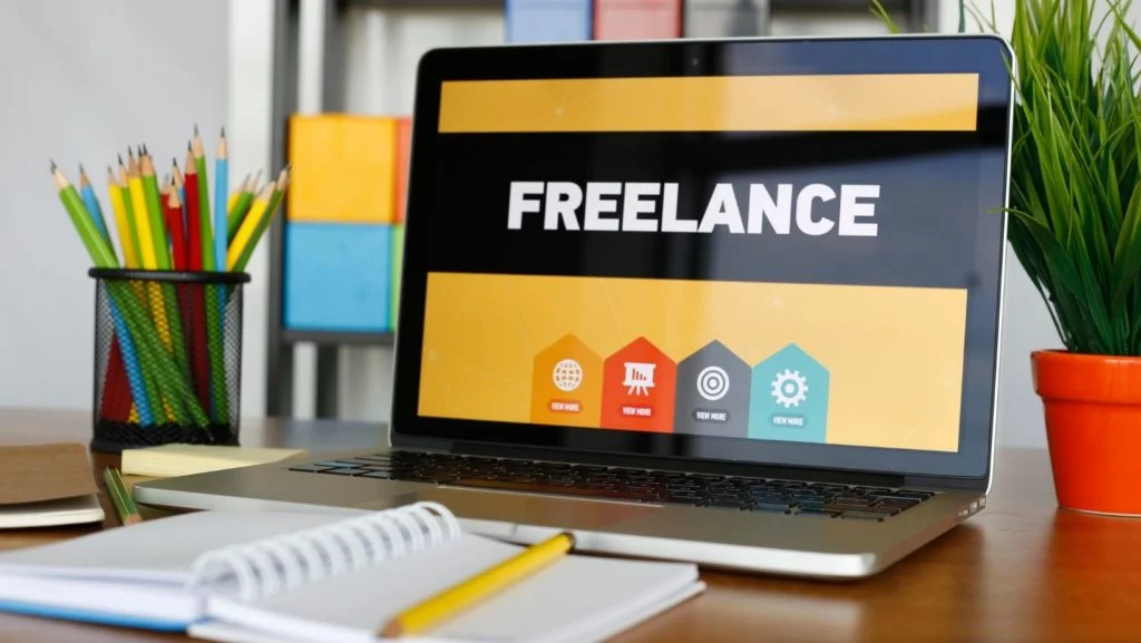 Laptop con "freelance" en la pantalla con un cuaderno en primer plano y lápices en segundo plano