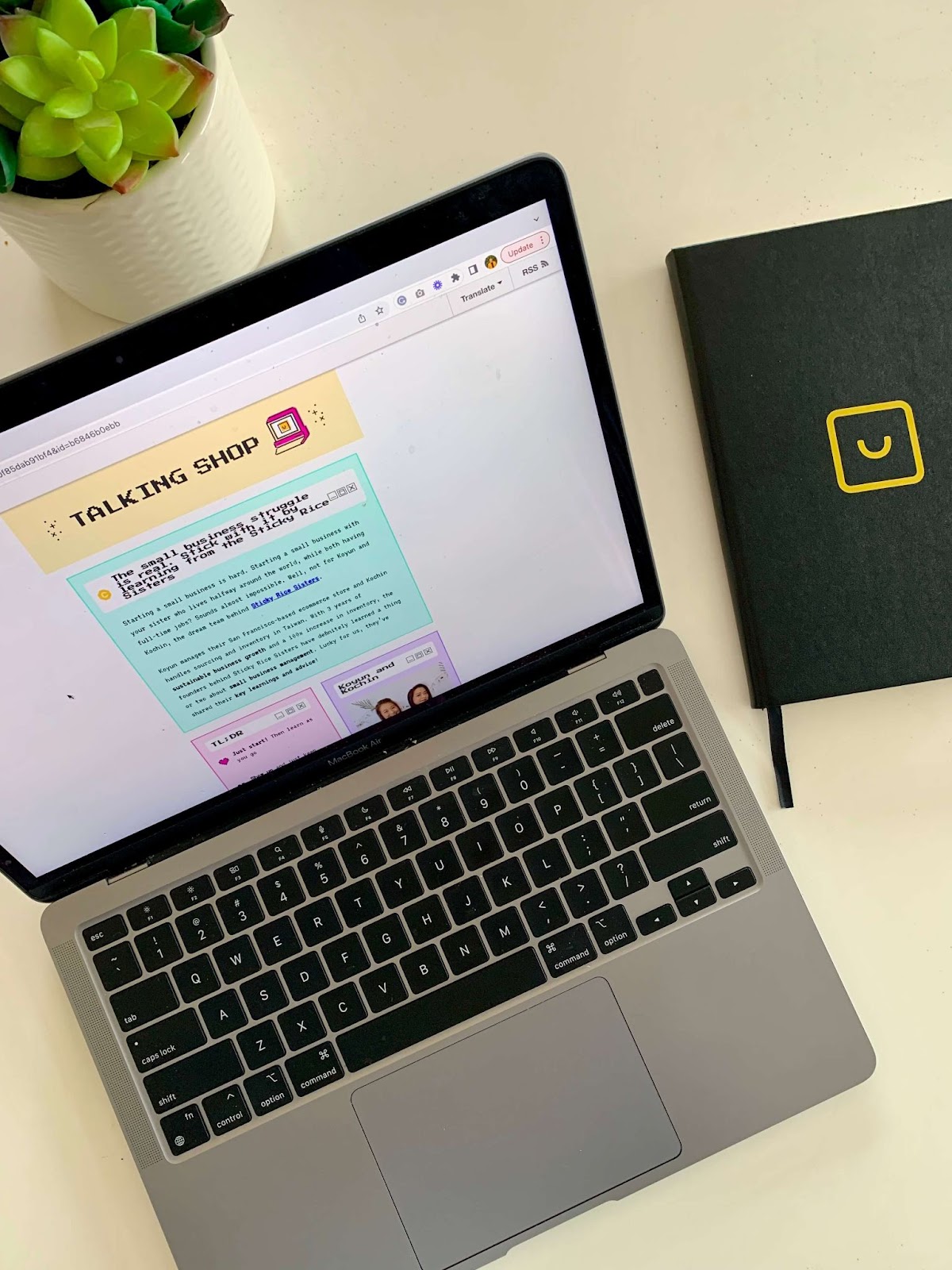 Talking Shop Smile Newsletter–Gambar MacBook di atas meja putih terbuka ke buletin Talking Shop, yang merupakan buletin gaya 8-bit dengan warna-warna cerah. Ada buku catatan hitam dengan logo senyum kuning di atasnya dan tanaman sukulen di atas meja.