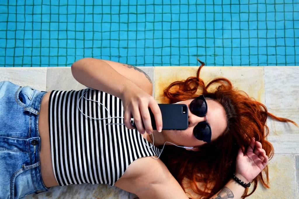 Mujer latina acostada junto a la piscina con gafas de sol y camiseta sin mangas mientras habla con su teléfono