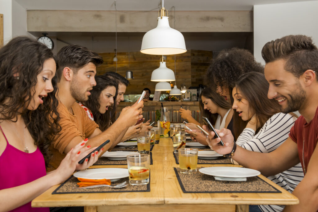 Ein Bild zeigt eine Gruppe von Menschen, die am selben Tisch sitzen und alle ihre Telefone benutzen.